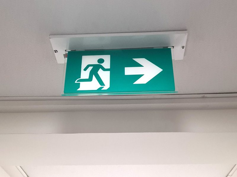 green exit sign at the ceiling in the narrow hall with regular ceiling lights white walls