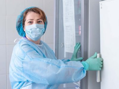 nurse in a medical cap, mask and blue protective gown opens the refrigerator door with the vaccine.