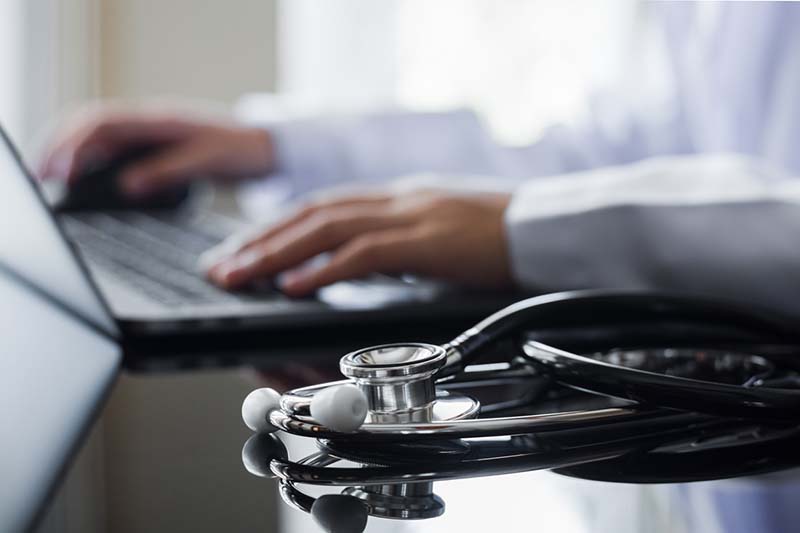 Female doctor in white lab coat typing on laptop computer with notebook and medical stethoscope on the desk at workplace. Medical technology ,Electronic health record system (EMR) concept