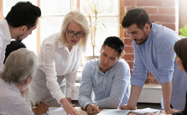 Serious old mature woman team leader coach teach young workers explain paper business plan at group meeting, focused senior female teacher mentor training diverse staff at corporate office workshop